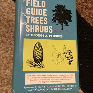 Könnte beinhalten: Ein blauer Buchumschlag mit dem Titel "A Field Guide to Trees and Shrubs" von George A. Petrides. Das Buch ist eine zweite Auflage und wird von der National Audubon Society und der National Wildlife Federation gefördert.