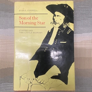 Può includere: Un libro vintage intitolato "Son of the Morning Star" di Evan S. Connell. La copertina presenta un'illustrazione in bianco e nero di un uomo in uniforme militare e cappello. Il sottotitolo del libro recita "Custer and the Little Bighorn."