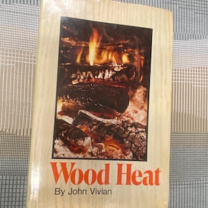 May include: A book titled "Wood Heat" by John Vivian. The cover features a photograph of a burning fire with orange and yellow flames. The book has a light wood-grain border.