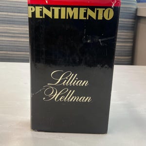 Puede incluir: Un libro de tapa dura titulado "Pentimento" de Lillian Hellman. El libro tiene una cubierta negra con letras doradas. La parte superior del libro es roja. El libro está de pie sobre una superficie clara.