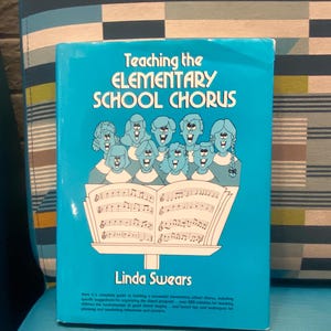 May include: A turquoise book titled "Teaching the Elementary School Chorus" by Linda Swears. The cover features an illustration of a choir singing from sheet music. The book is on a blue chair.