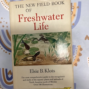 May include: The cover of a book titled "The New Field Book of Freshwater Life" by Elsie B. Klots. The cover features an illustration of a frog, a turtle, and a dragonfly in a pond setting. The book is a guide to the recognition and study of aquatic plants and animals in North America. It includes over 700 illustrations.