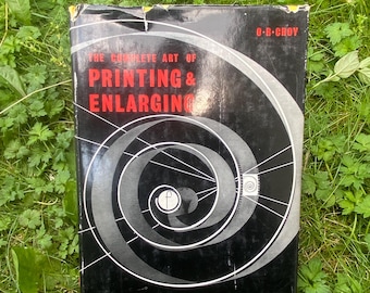 Puede incluir: Un libro antiguo titulado "The Complete Art of Printing & Enlarging" de O.R. Croy. El libro tiene una cubierta negra con un diseño en espiral plateado y texto rojo. El libro está sobre hierba verde.