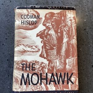 May include: A vintage book titled "The Mohawk" by Codman Hislop, illustrated by Letterio Calapai. The cover features a brown and white illustration of a Native American figure and landscape. The book is part of the "Rivers of America" series.