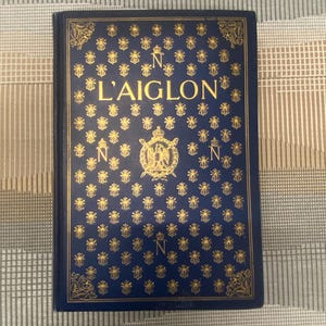 Könnte beinhalten: Ein Hardcover-Buch mit dem Titel "L'Aiglon" und einem marineblauen Einband. Der Einband ist mit goldenen Bienen, dem Buchstaben "N" und goldenen Verzierungen versehen. Der Titel ist in Goldbuchstaben gehalten.