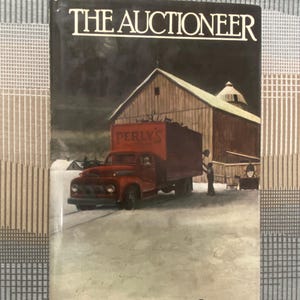 May include: A book cover for "The Auctioneer" by Joan Samson. The cover features a red truck with a large cargo bed parked in front of a wooden barn. The title is in white block letters, and the author's name is at the bottom.