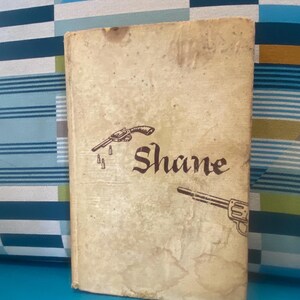 May include: A vintage book titled "Shane" with a tan cover. The cover features illustrations of revolvers and bullet casings. The title is in a stylized font. The book shows signs of age and wear.