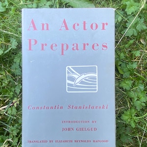 Può includere: Libro grigio intitolato "An Actor Prepares" di Constantin Stanislavski, con testo rosso e grafica bianca. Il libro è sull'erba verde. Introduzione di John Gielgud.