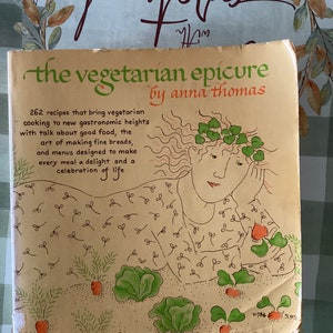 May include: A vintage cookbook titled "The Vegetarian Epicure" by Anna Thomas. The cover features a hand-drawn illustration of a woman with a crown of leaves, surrounded by carrots and leafy greens. The text "262 recipes that bring vegetarian cooking to new gastronomic heights" is printed on the cover.