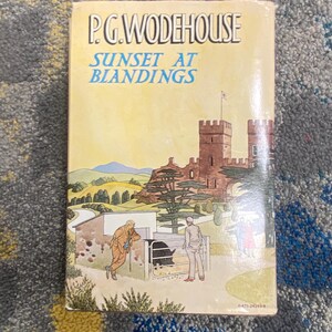 May include: A book titled Sunset at Blandings by P.G. Wodehouse with a cover illustration featuring a large stone castle, rolling green hills, and two figures standing near a pig pen containing a black pig. The text is printed in black and blue.