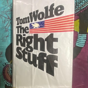 May include: A paperback book titled "The Right Stuff" by Tom Wolfe. The cover is white with black text and a stylized American flag graphic. The flag features red and white stripes and a blue field with a white eagle.