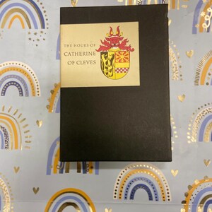 May include: A black book with a gold label that reads "The Hours of Catherine of Cleves". The label features a coat of arms with a red dragon and a yellow and black shield.