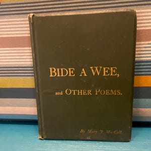 Peut inclure: Un livre vintage vert foncé intitulé "Bide a Wee, and Other Poems" avec des lettres dorées. Le livre est de Mary J. McColl. Le livre est sur une surface bleue avec un fond rayé coloré.