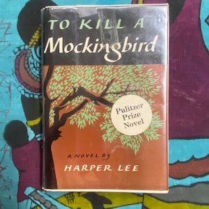 Puede incluir: Un ejemplar de tapa dura de "To Kill a Mockingbird" de Harper Lee. La portada del libro presenta una ilustración de un árbol con hojas verdes y un tronco marrón sobre un fondo naranja. El título está en blanco y se ve una pegatina de "Pulitzer Prize Novel".
