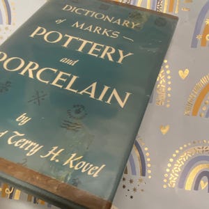 Pode incluir: Um livro turquesa intitulado "Dictionary of Marks - Pottery and Porcelain" de Terry H. Kovel. O título está em letras douradas. O livro está sobre um fundo azul claro com padrões de arco-íris e corações.