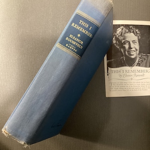 May include: A blue hardcover book titled "This I Remember" by Eleanor Roosevelt. The book is slightly worn and has a white label on the spine. The book is lying on a brown surface.