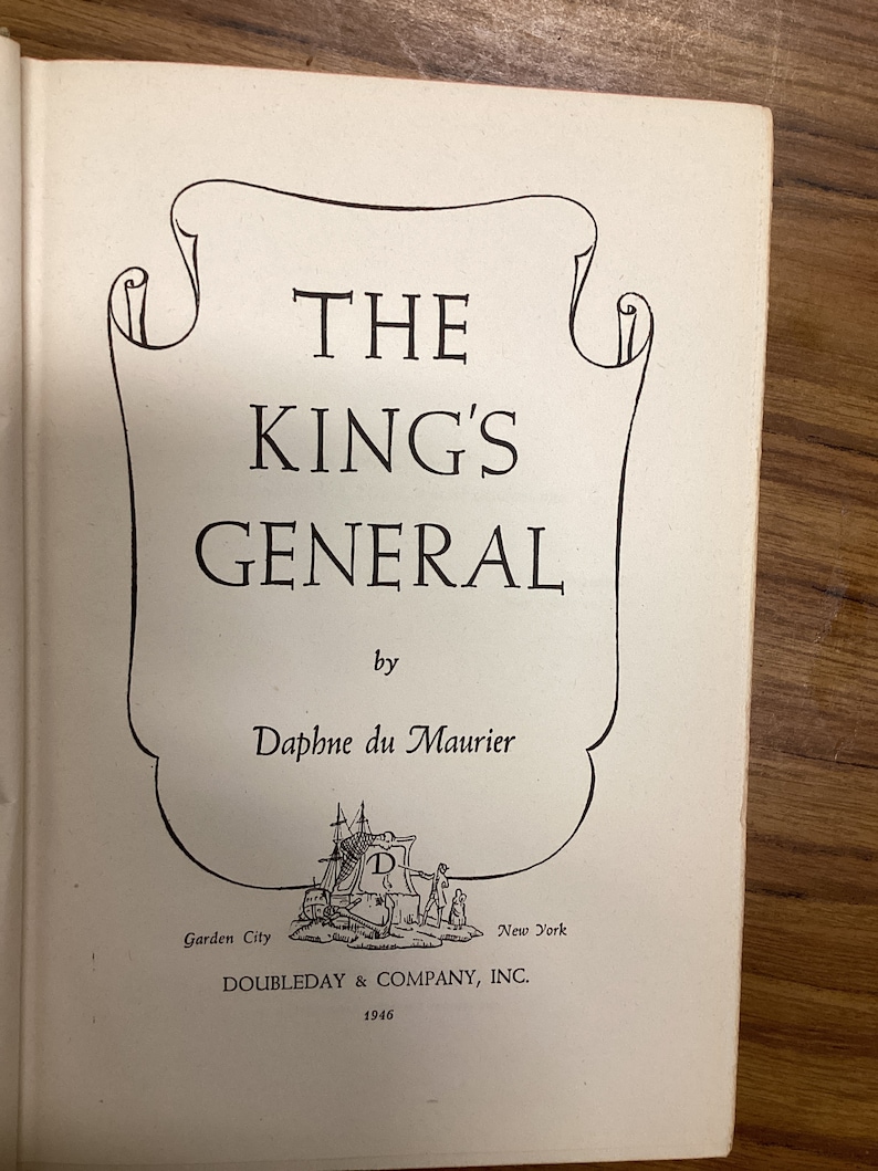 以下が含まれることがあります： ダフネ・デュ・モーリア著「THE KING'S GENERAL」のタイトルが書かれた開いた本のページ。タイトルは太字のセリフ体で、装飾的なフレーム内にあります。「DOUBLEDAY & COMPANY, INC.」と年号「1946」を含む出版社の情報も表示されています。