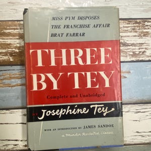 Puede incluir: Un libro antiguo titulado "Three by Tey" de Josephine Tey. El libro tiene una cubierta gris con detalles en rojo y negro. El título está en letras blancas grandes. El libro también incluye los títulos "Miss Pym Disposes", "The Franchise Affair" y "Brat Farrar".