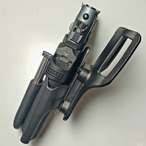 K&ouml;nnte beinhalten: Eine schwarze Pistole in einer schwarzen Holster. Die Pistole hat einen grauen Schlitten und eine Digitalanzeige. Die Holster hat eine G&uuml;rtelschlaufe. Das Bild ist aus der Vogelperspektive aufgenommen.