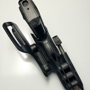 K&ouml;nnte beinhalten: Schwarze Polymer-Pistolenholster mit G&uuml;rtelschlaufe und Magazinhalter. Das Holster ist so konzipiert, dass es eine Schusswaffe sicher h&auml;lt. Das Design umfasst ein R&uuml;ckhaltesystem und besteht aus strapazierf&auml;higem, geformtem Kunststoff.