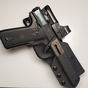 K&ouml;nnte beinhalten: Eine schwarze Schusswaffe mit silbernem Lauf und einem Rotpunktvisier, gesichert in einer schwarzen Halterung. Ein Magazin ist in die Halterung eingesetzt. Die Schusswaffe liegt auf einer wei&szlig;en Oberfl&auml;che.