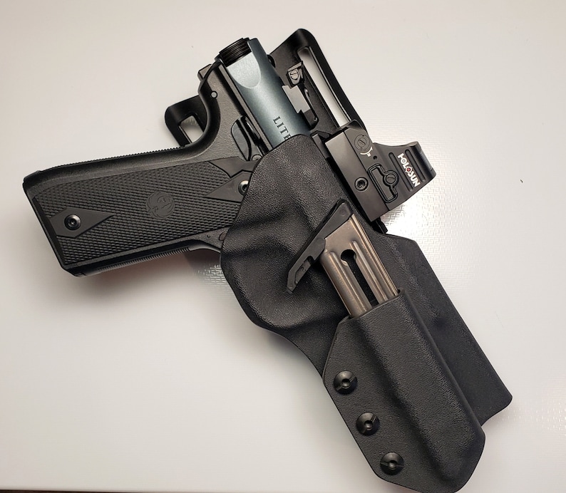 K&ouml;nnte beinhalten: Eine schwarze Schusswaffe mit silbernem Lauf und einem Rotpunktvisier, gesichert in einer schwarzen Halterung. Ein Magazin ist in die Halterung eingesetzt. Die Schusswaffe liegt auf einer wei&szlig;en Oberfl&auml;che.