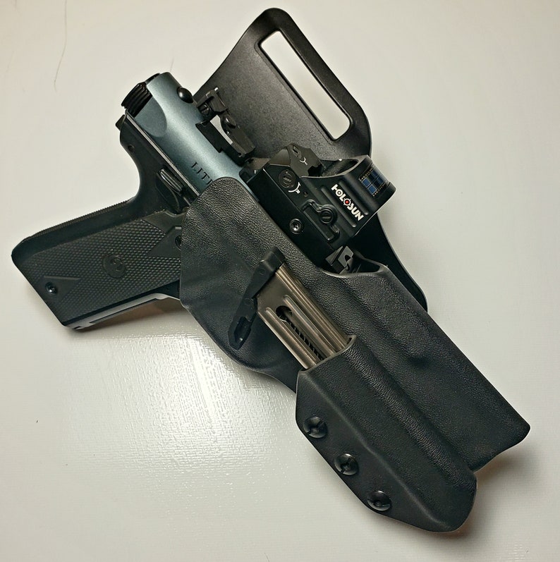 K&ouml;nnte beinhalten: Eine schwarze Polymerholster mit einer halbautomatischen Pistole und einem Magazin. Die Pistole hat einen grauen Schlitten und ein Rotpunktvisier. Die Holster ist f&uuml;r das Tragen am G&uuml;rtel konzipiert.