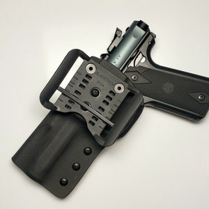K&ouml;nnte beinhalten: Eine schwarze Schusswaffe mit silbernem Schlitten ist teilweise in einer schwarzen Holster eingesetzt. Das Holster hat eine G&uuml;rtelschlaufe und ist mit "Bladetech U.S.A." gekennzeichnet. Die Waffe hat einen strukturierten Griff vor wei&szlig;em Hintergrund.