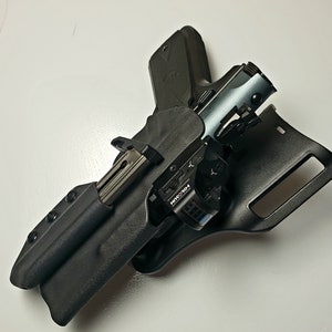 K&ouml;nnte beinhalten: Eine schwarze Holster mit einer silbernen und schwarzen Handfeuerwaffe und einem silbernen Magazin. Die Handfeuerwaffe hat ein Rotpunktvisier. Das Holster besteht aus einem strapazierf&auml;higen Material.