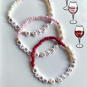 Könnte beinhalten: Drei Perlenarmbänder mit den Worten "Wine o'clock" in goldenen Buchstaben auf weißen Perlen. Die Armbänder sind übereinander gelegt, wobei das obere Armband weiß, das mittlere Armband rosa und das untere Armband rot ist. Die Armbänder sind mit weißen Perlen verziert.