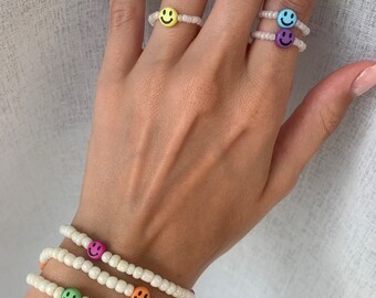 May include: A set of three white beaded bracelets with colourful smiley face charms. The bracelets are stacked on a person's wrist.