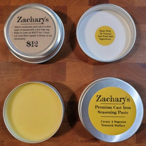 May include: Two silver tins with yellow labels. The top tin has a label that reads "Zachary's" and includes instructions for using the seasoning paste. The bottom tin has a label that reads "Zachary's Premium Cast Iron Seasoning Paste" and includes the text "Create A Superior Nonstick Surface".