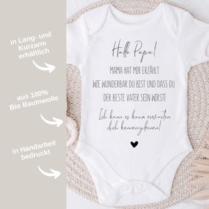 May include: White baby onesie with black text that reads "Hallo Papa! Mama hat mir erz&auml;hlt wie wunderbar du bist und dass du der beste Vater sein wirst! Ich kann es kaum erwarten dich kennenzulernen!"  The onesie is available in long and short sleeves and is made from 100% organic cotton.