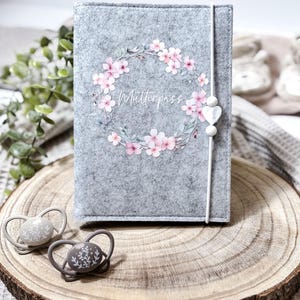 Könnte beinhalten: Ein grauer Filzbezug mit dem Wort "Mutterpass" in Schrift, umgeben von einem Kranz aus rosa Blüten. Ein weißes Band mit einer herzförmigen Perle sichert den Bezug. Zwei Schnuller mit dekorativen Designs liegen auf einer Holzscheibe.
