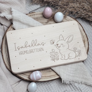 Könnte beinhalten: Helles Holzschneidebrett mit dem Namen "Isabellas" und dem Text "KRÜMELBRETTCHEN" eingraviert. Ein lächelnder Hase und ein Schmetterling sind ebenfalls eingraviert. Dekorative Ostereier umgeben das Brett.