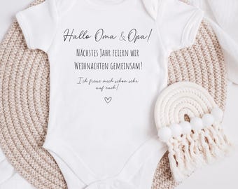 May include: White baby onesie with black text that reads "Hallo Oma & Opa! Nächstes Jahr feiern wir Weihnachten gemeinsam! Ich freue mich schon sehr auf euch!" with a small heart symbol.