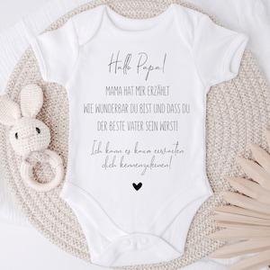 May include: White baby onesie with black text that reads "Hallo Papa! Mama hat mir erz&auml;hlt wie wunderbar du bist und dass du der beste Vater sein wirst! Ich kann es kaum erwarten dich kennenzulernen!" with a small black heart at the bottom.