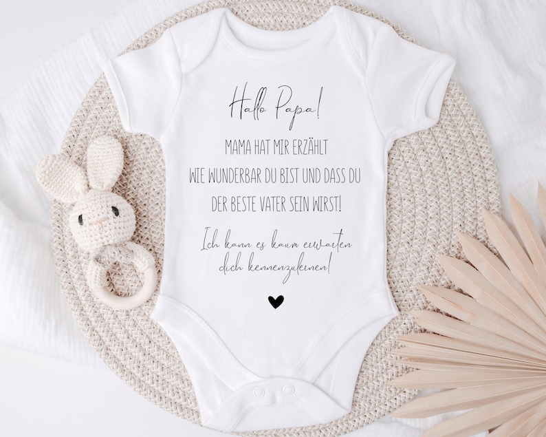 May include: White baby onesie with black text that reads "Hallo Papa! Mama hat mir erz&auml;hlt wie wunderbar du bist und dass du der beste Vater sein wirst! Ich kann es kaum erwarten dich kennenzulernen!" with a small black heart at the bottom.