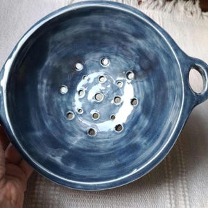Può includere: Scolapasta in ceramica blu fatto a mano, con due manici. Lo scolapasta ha una finitura lucida e numerosi fori di drenaggio sul fondo. La ciotola è rotonda e sembra robusta, adatta per l'uso in cucina.