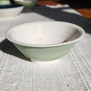 Può includere: Una ciotola in ceramica fatta a mano con esterno verde chiaro e interno bianco maculato. La ciotola ha una forma arrotondata e una piccola base bianca. La ciotola è su una superficie bianca strutturata.