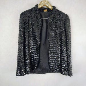 May include: A black sequin jacket with a collar, hanging on a gold glitter hanger. The jacket is open, revealing a black lining. The sequins create a shimmering effect, reflecting light. This vintage jacket is a statement piece.