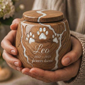 Puede incluir: Una urna redonda de madera para mascotas con acabado de veta natural y una incrustación de resina blanca en el centro. La urna está grabada con tres huellas de patas, el nombre Leo, los años 2012-2019 y la frase forever loved.