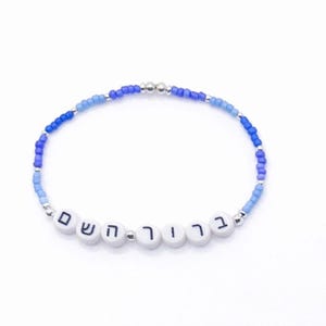 Può includere: Un braccialetto di perline con perline blu e argento. Il braccialetto presenta perline di lettere bianche che compongono il testo ebraico. Le perline sono infilate su un cordino flessibile, creando una forma circolare.