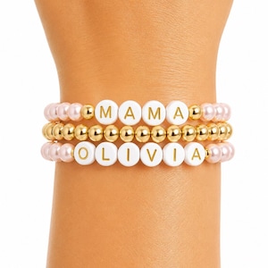 May include: A stack of three beaded bracelets. The top bracelet features light pink pearls and white round beads with the gold text MAMA. The middle bracelet consists of small gold metallic beads. The bottom bracelet features light pink pearls and white round beads with the gold text OLIVIA.