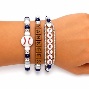 Può includere: Tre braccialetti di perline con un ciondolo a forma di baseball, perline quadrate marroni che compongono la scritta YANKEES e una catenina argentata con perline bianche rotonde che compongono YANKEES in lettere rosse, con perline blu, bianche e argento.