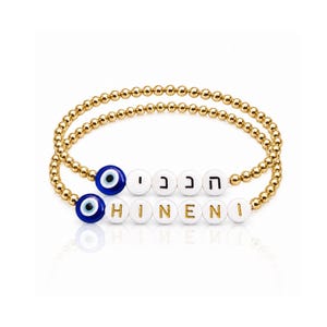 May include: Two gold beaded bracelets. One bracelet features white letter beads spelling "HINENI" and Hebrew letters. The other bracelet has white letter beads and two blue evil eye beads.
