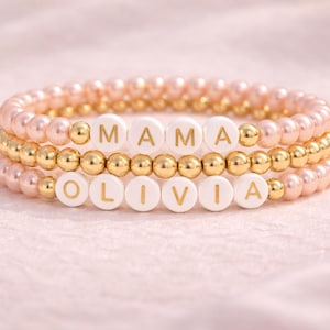 Pode incluir: Três pulseiras de contas. As pulseiras superior e inferior são feitas de contas rosa. A pulseira do meio é feita de contas douradas. A pulseira superior diz "MAMA" e a pulseira inferior diz "OLIVIA" com contas de letras brancas.