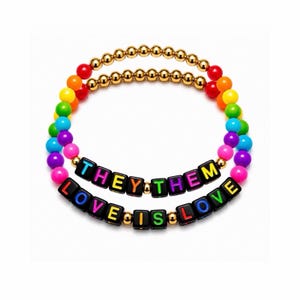 May include: Two stacked beaded bracelets featuring metallic gold beads and vibrant rainbow-colored round beads. One bracelet displays black square letter beads spelling THEY THEM, and the other displays black square letter beads spelling LOVE IS LOVE.