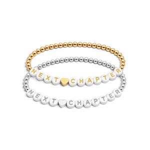 May include: Two beaded bracelets featuring white round beads with gold and silver lettering that spells out NEXT CHAPTER. One bracelet includes a gold heart bead, while the other features a silver heart bead. The bands are made of metallic beads.