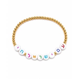 May include: Gold beaded bracelet with white letter beads spelling out Hebrew text in various colors. The bracelet has a gold chain and is set against a white background.